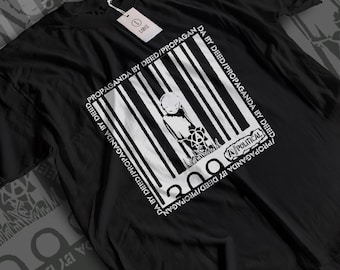 May include: A black short-sleeve t-shirt featuring a white graphic print of a figure standing behind vertical bars resembling a barcode. The text "PROPAGANDA BY DEED" repeats around the square border, with "A-POLITICAL" printed in the corner.