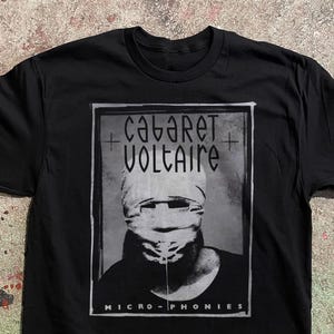 May include: Black t-shirt featuring a grayscale image of a person with a wrapped head, framed by the words "Cabaret Voltaire" and "Micro-Phonies" in a stylized font. The design is centered on the shirt.