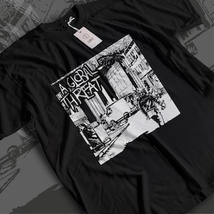 Könnte beinhalten: Ein schwarzes Kurzarm-T-Shirt mit einem weißen Grafikdruck einer Stadtstraßenszene mit einer Person, einem Lieferwagen und dem Text A GLOBAL THREAT. Ein weißes rechteckiges Etikett am Kragen zeigt den Buchstaben L und das Wort LARGE.
