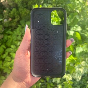 Puede incluir: Funda de tel&eacute;fono negra con un patr&oacute;n geom&eacute;trico en el interior. La funda tiene un recorte para la c&aacute;mara y se sostiene contra un fondo de follaje verde. La funda est&aacute; dise&ntilde;ada para proteger un tel&eacute;fono inteligente.