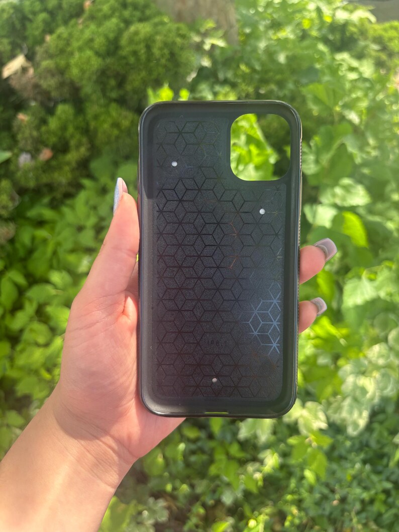 Puede incluir: Funda de tel&eacute;fono negra con un patr&oacute;n geom&eacute;trico en el interior. La funda tiene un recorte para la c&aacute;mara y se sostiene contra un fondo de follaje verde. La funda est&aacute; dise&ntilde;ada para proteger un tel&eacute;fono inteligente.