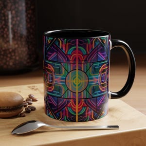 Op de afbeelding: Een koffiemok met een zwart handvat en een levendig, geometrisch ontwerp in paars, groen, blauw en geel. De binnenkant van de mok is zwart. De mok staat op een houten oppervlak met een lepel, een macaron en koffiebonen.