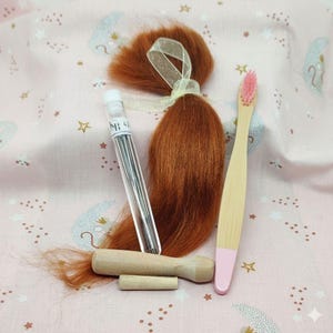 Peut inclure: Ensemble d'objets comprenant une mèche de cheveux roux attachée avec un ruban, une fiole transparente d'aiguilles étiquetée "NI 42", une brosse à dents en bois avec une pointe rose et un outil en bois. Les articles sont disposés sur un tissu rose à motifs célestes.