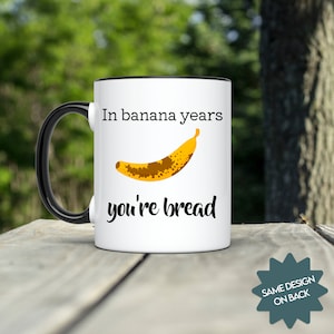 Può includere: Tazza in ceramica bianca con bordo e manico neri. La tazza ha una grafica nera che dice "In banana years you're bread" con una banana di cartone animato con macchie marroni.