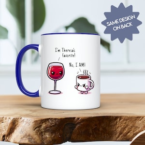 Könnte beinhalten: Weiße Keramik-Kaffeetasse mit blauem Rand. Die Tasse zeigt eine Comic-Illustration eines Glases Rotwein und einer Kaffeetasse mit Gesichtern. Der Text auf der Tasse lautet "Ich bin Theresas Favorit! Nein, ich bin es!"