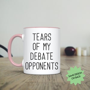 May include: White ceramic coffee mug with a pink rim and handle. The mug has black text that reads "Tears of my Debate Opponents".