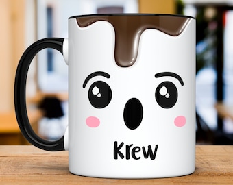 Könnte beinhalten: Weiße Keramiktasse mit schwarzem Henkel. Die Tasse hat ein braunes, schokoladenartiges Design, das an der Vorderseite heruntertropft. Die Tasse hat ein Comic-Gesicht mit großen Augen, einem überraschten Ausdruck und dem Namen "Krew" unter dem Gesicht.