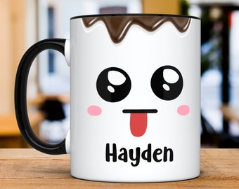 Könnte beinhalten: Eine weiße Keramiktasse mit schwarzem Henkel und schwarzem Rand. Die Tasse hat ein Comic-Gesicht mit großen Augen, einer herausgestreckten Zunge und rosa Wangen. Die Tasse hat ein braunes, schokoladenartiges Design am Rand und den Namen "Hayden" in Schwarz unter dem Gesicht.