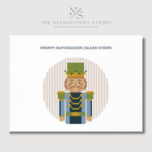 May include: A needlepoint pattern of a preppy nutcracker with a green hat, gold crown, and blue and black uniform. The pattern is called "Preppy Nutcracker | Blush Stripe".
