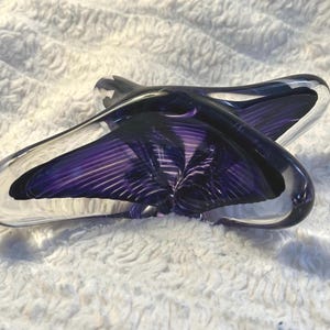 May include: A clear and purple art glass sculpture with a unique, abstract shape. The sculpture features a deep purple interior with linear striations, encased in clear glass. The design is reminiscent of a stylized flower or star.