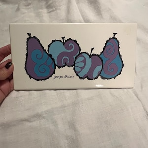 May include: A rectangular ceramic tile with a white background. The tile features a stylized illustration of pears and apples in shades of purple and blue, outlined in black. The text "Georges Briard" is printed in black.
