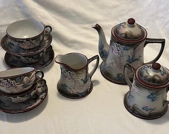 Könnte beinhalten: Ein vintage japanisches Teeservice mit Drachenmotiv. Das Set besteht aus einer Teekanne, einem Milchkännchen, einer Zuckerdose und zwei Teetassen mit Untertassen. Das Porzellan ist mit einem weißen Drachen auf grau-braunem Hintergrund verziert.