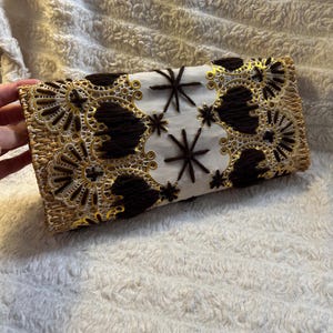 May include: A rectangular clutch bag featuring a woven straw base, a central white mesh panel, and dark brown yarn embroidery. The design includes gold-toned lace-like floral patterns and star-shaped stitching accents across the front surface.