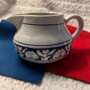 May include: A small, white ceramic creamer with a blue rabbit and floral design. The creamer has a handle and a spout. The exterior has a crackle glaze. The creamer is approximately 4 inches tall.