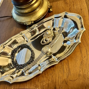 May include: An oval silver-toned metal tray with a scalloped edge containing a dark beaded bracelet, a silver-toned link watch with a rectangular face, and a circular brooch featuring a central white pearl surrounded by small clear stones.