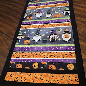 May include: A Halloween table runner with a black border and multiple stripes of fabric featuring gnomes, pumpkins, and stars. The fabric is in shades of purple, green, black, orange, and white.