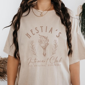 May include: A beige t-shirt with a graphic design that says "Hestia's Introvert Club The Original Homebody". The graphic features a torch with flames and a wreath of leaves.