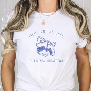 May include: White t-shirt with a blue raccoon graphic and the text "Livin' on the edge of a mental breakdown".
