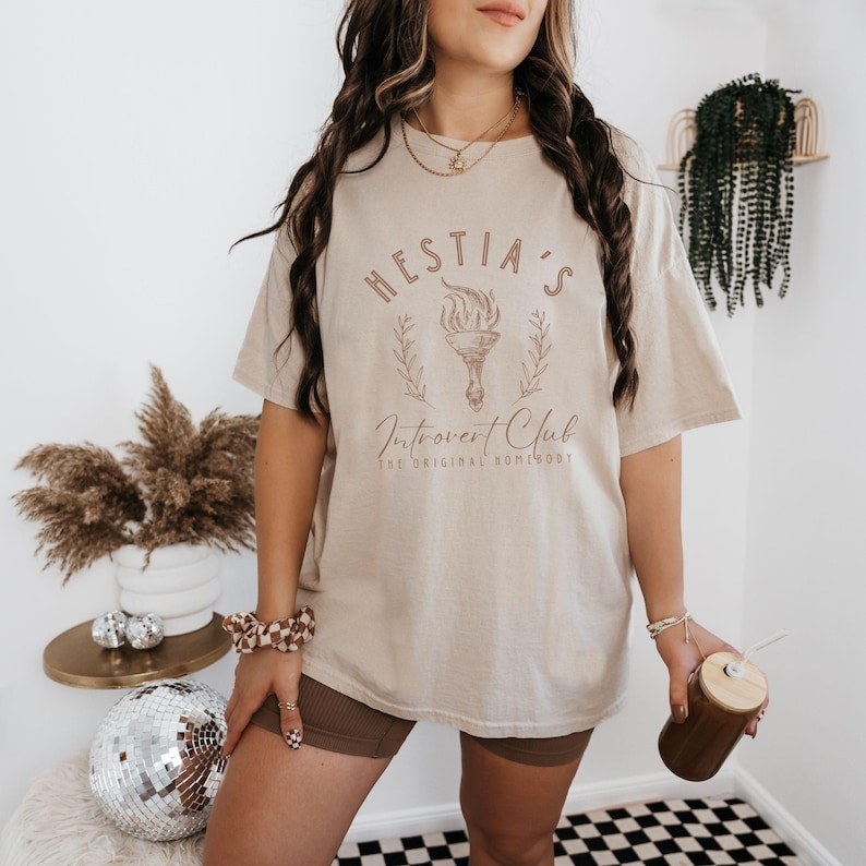 May include: A beige t-shirt with a graphic design that says "Hestia's Introvert Club The Original Homebody". The graphic features a torch with flames and a wreath of leaves.