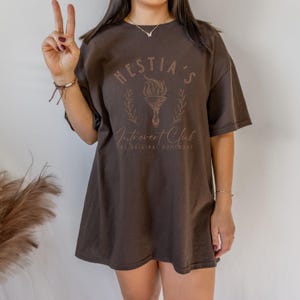 May include: A brown oversized t-shirt with a graphic design that says "Hestia's Introvert Club The Original Homebody". The graphic features a stylized torch with flames and a floral design.