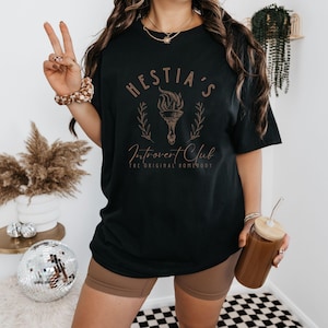 May include: Black t-shirt with a gold graphic that says "Hestia's Introvert Club The Original Homebody".