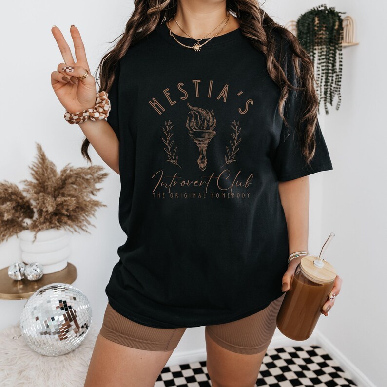 May include: Black t-shirt with a gold graphic that says "Hestia's Introvert Club The Original Homebody".