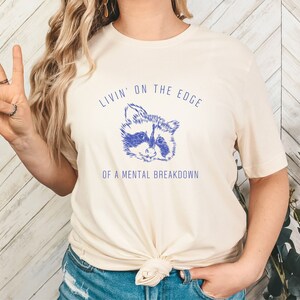 May include: A cream colored t-shirt with a blue raccoon graphic and the text "Livin' on the edge of a mental breakdown".
