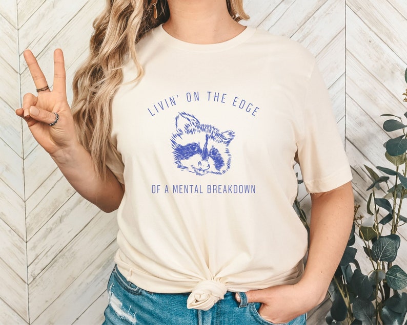 May include: A cream colored t-shirt with a blue raccoon graphic and the text "Livin' on the edge of a mental breakdown".