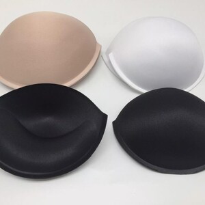 Op de afbeelding: Vier bh-inlegstukken in verschillende kleuren: beige, wit, zwart en zwart met een naad in het midden. De inlegstukken zijn gemaakt van schuim en zijn ontworpen om volume aan de borst toe te voegen.