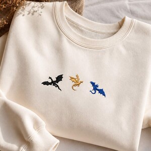 Op de afbeelding: Cr&egrave;mekleurige sweatshirt met geborduurde draken in zwart, goud en blauw. De draken zijn gestileerd en lijken te vliegen. De sweatshirt heeft een ronde hals en lange mouwen, ideaal voor vrijetijdskleding.