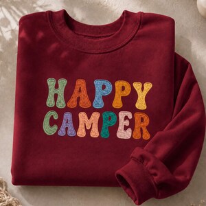 Puede incluir: Una sudadera de cuello redondo de color rojo oscuro con el texto HAPPY CAMPER bordado en una fuente ondulada multicolor. Las letras están cosidas en tonos verde, naranja, azul, amarillo, morado y rosa, creando un contraste con la tela.