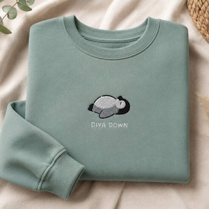 May include: A sage green crewneck sweatshirt featuring an embroidered graphic of a penguin lying on its stomach. Below the penguin, the text DIVA DOWN is embroidered in white thread. The sweatshirt has ribbed cuffs and a ribbed neckline.