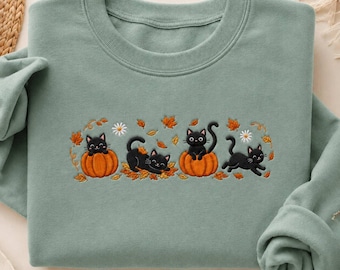 May include: A sage green crewneck sweatshirt featuring an embroidered design of four black cats playing with orange pumpkins and scattered autumn leaves, accented by small white daisy flowers across the centre of the chest.