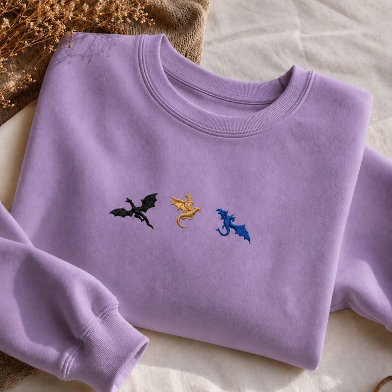Op de afbeelding: Een lila sweatshirt met geborduurde draken in zwart, goud en blauw. De crewneck sweatshirt heeft lange mouwen en geribde manchetten en kraag. De vliegende draken geven het kledingstuk een grillige uitstraling.