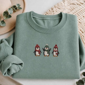 Könnte beinhalten: Ein salbeigrünes Sweatshirt mit Rundhalsausschnitt und einer Stickerei von drei nebeneinander stehenden Pinguinen. Jeder Pinguin trägt eine kleine Strickmütze und einen Schal in Rot- und Grüntönen. Das Sweatshirt hat lange Ärmel und einen Rippkragen.
