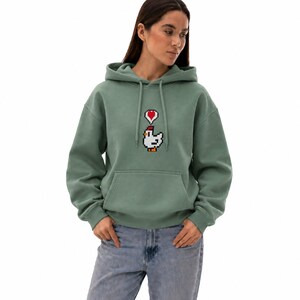 May include: Sage green hoodie with a pixelated chicken graphic and a red heart above it. The hoodie has a drawstring hood and a front pocket. The model is wearing blue jeans.
