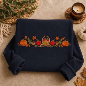 Pode incluir: Uma camisola azul-marinho escuro com um bordado horizontal central que apresenta um cesto entrançado cheio de maçãs vermelhas, ladeado por abóboras cor de laranja, bolotas castanhas, folhas verdes e pequenas bagas vermelhas.