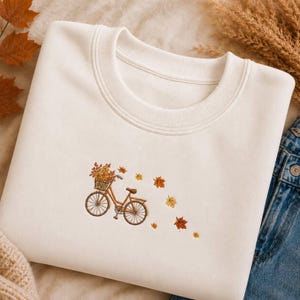 Op de afbeelding: Een crèmekleurige sweater met ronde hals en een kleine, gecentreerde borduursel van een bruine fiets met een mand vol herfstbladeren. Verschillende oranje, gele en bruine herfstbladeren zijn achter de fiets geborduurd.
