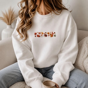 Könnte beinhalten: Ein weißes Sweatshirt mit Rundhalsausschnitt und einem mittigen, horizontalen Grafikdruck von Waldmotiven, darunter ein Fuchs, ein Igel, ein Eichhörnchen, Pilze, Eicheln und Herbstblätter in den Farben Orange, Braun und Rot, kombiniert mit blauen Jeans.