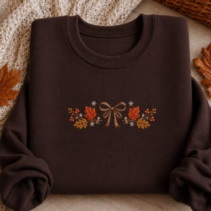 Peut inclure: Un sweat-shirt à col rond marron foncé orné d'une broderie centrale représentant un nœud ruban marron entouré de feuilles d'érable orange et jaunes, de petites fleurs blanches en forme d'étoile et de grappes de baies rouges.