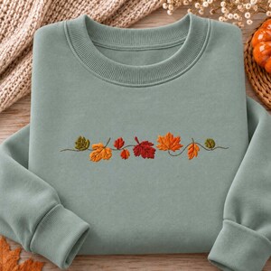 Peut inclure: Un sweat-shirt à col rond vert sauge orné d'une broderie horizontale centrale représentant diverses feuilles d'automne dans des tons orange, rouge, jaune et vert olive, reliées par une fine tige brune incurvée sur la poitrine du vêtement.
