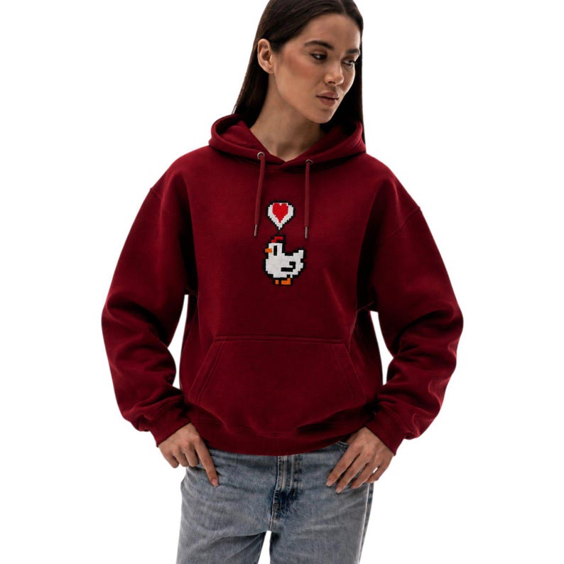 May include: A maroon hoodie with a pixelated white chicken graphic and a red heart above it. The hoodie has a drawstring hood and a front pocket. The sleeves are long, and the material appears to be a soft fabric.