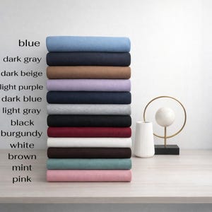 Op de afbeelding: Een stapel gevouwen stoffen in verschillende kleuren, waaronder blauw, donkergrijs, donkerbeige, lichtpaars, donkerblauw, lichtgrijs, zwart, bordeauxrood, wit, bruin, mint en roze. Kleur labels zijn zichtbaar.
