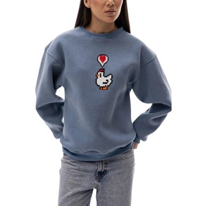 May include: A blue sweatshirt with a pixelated chicken graphic and a red heart above it. The sweatshirt has long sleeves and a crew neck. The model is wearing blue jeans.