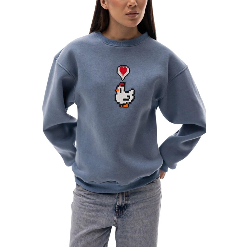 May include: A blue sweatshirt with a pixelated chicken graphic and a red heart above it. The sweatshirt has long sleeves and a crew neck. The model is wearing blue jeans.