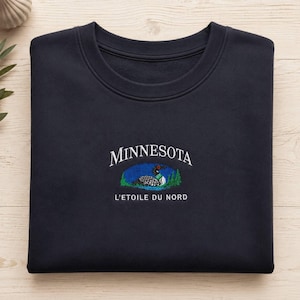Puede incluir: Una sudadera de cuello redondo en color azul marino oscuro con un diseño bordado en el pecho. El bordado incluye el texto blanco MINNESOTA, un gráfico colorido de un colimbo en un lago con árboles verdes y el texto blanco L'ETOILE DU NORD.
