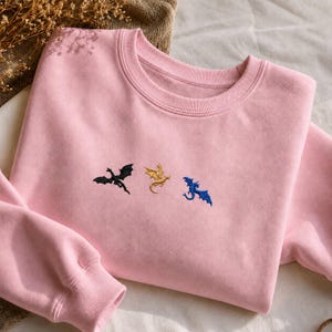 Op de afbeelding: Een lichtroze sweatshirt met geborduurde draken in zwart, goud en blauw. De draken zijn horizontaal over de borst geplaatst. De sweatshirt heeft een ronde hals en lange mouwen.