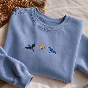 Op de afbeelding: Een lichtblauwe sweatshirt met geborduurde draken in zwart, goud en blauw. De crew neck sweatshirt is gemaakt van een zacht materiaal en heeft lange mouwen. De draken zijn op de borst geborduurd.
