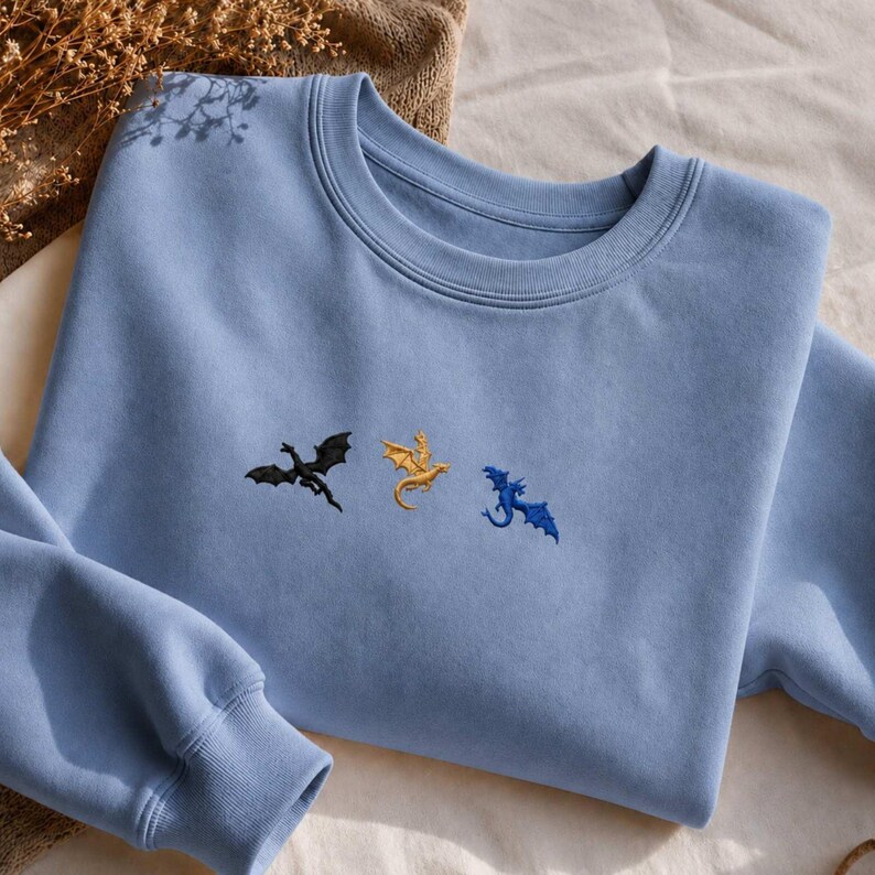 Op de afbeelding: Een lichtblauwe sweatshirt met geborduurde draken in zwart, goud en blauw. De crew neck sweatshirt is gemaakt van een zacht materiaal en heeft lange mouwen. De draken zijn op de borst geborduurd.