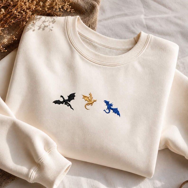 Op de afbeelding: Cr&egrave;mekleurige sweatshirt met geborduurde draken in zwart, goud en blauw. De draken zijn gestileerd en lijken te vliegen. De sweatshirt heeft een ronde hals en lange mouwen, ideaal voor vrijetijdskleding.